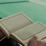 Nahaufnahme von einem aufgeschlagenen Koran in den Händen eines Mannes, auf einem Teppich sitzend.