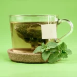 Grüner Tee mit Teebeutel vor einem hell-grünen Hintergrund auf einem Untersetzer in braun.
