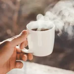 Eine Hand hält einen dampfenden schwarzen keto Kaffee in einer weißen Tasse.