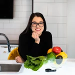 Frau mit schwarzem Pullover und schwarzer Brille lehnt sich in der Küche auf die Arbeitsfläche und schaut in die Kamera als Zeichen für Ernährungsberater Gehalt