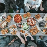 Ein Holztisch von oben mit Menschen sitzen und essen. Auf dem Tisch stehen viele Speisen, wie Burger, Pizza, Pommes als Zeichen für Omnivore Ernährung.